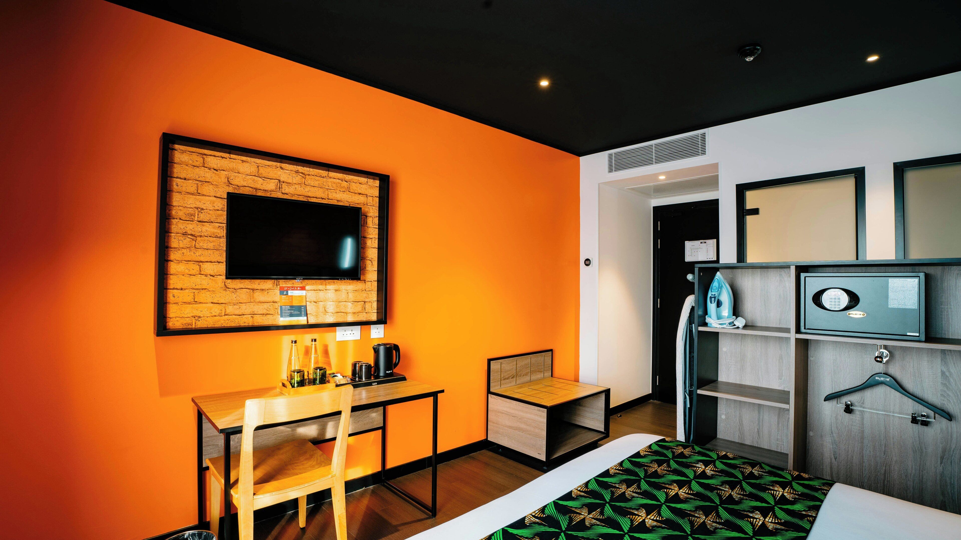A hotel room with a brick accent wall, a flat-screen TV, a desk with a chair, a bed with a patterned bedspread, and a wardrobe with a hanging iron.