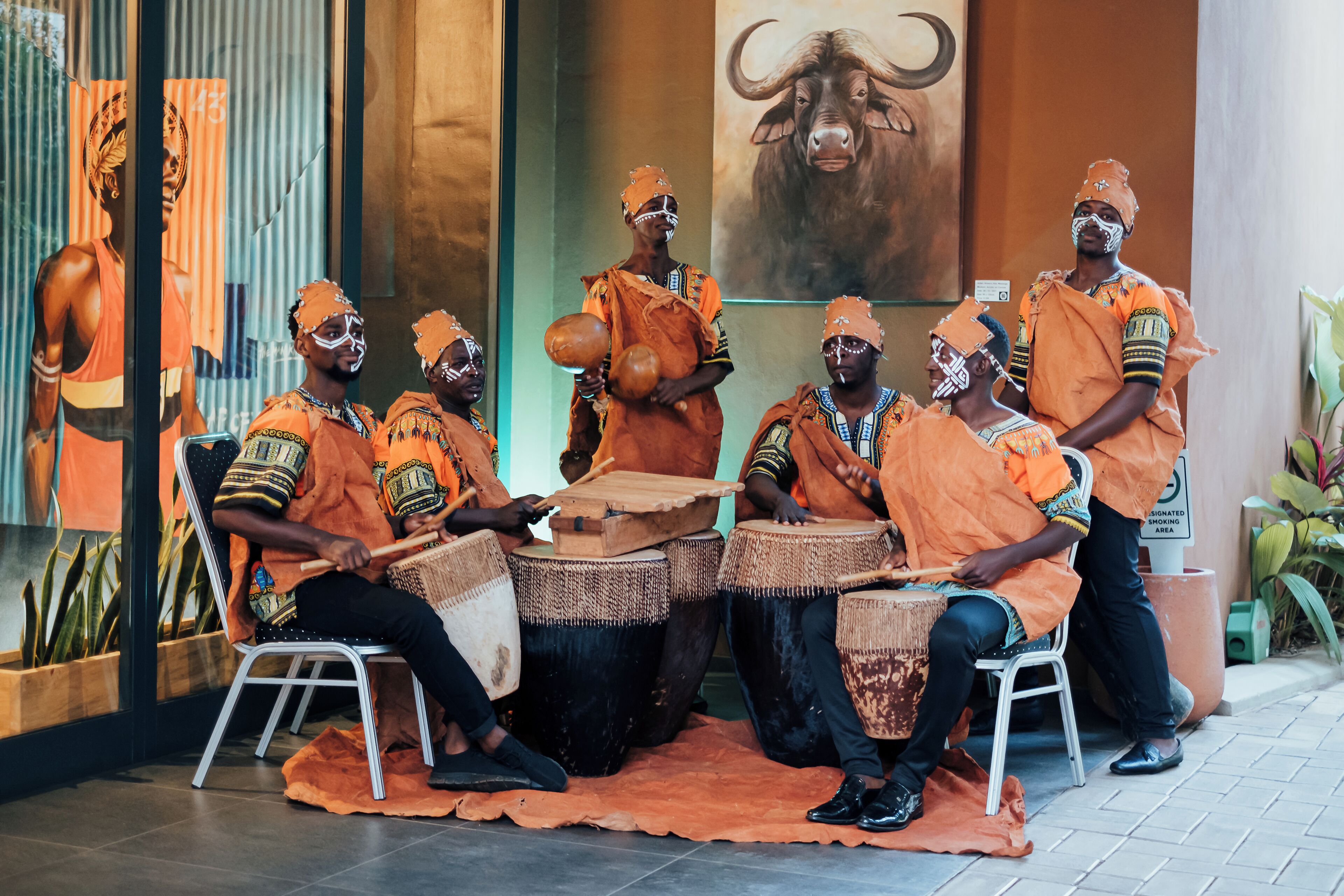 A group of people in traditional attire seated on chairs, playing drums in a room with a bull painting on the wall.