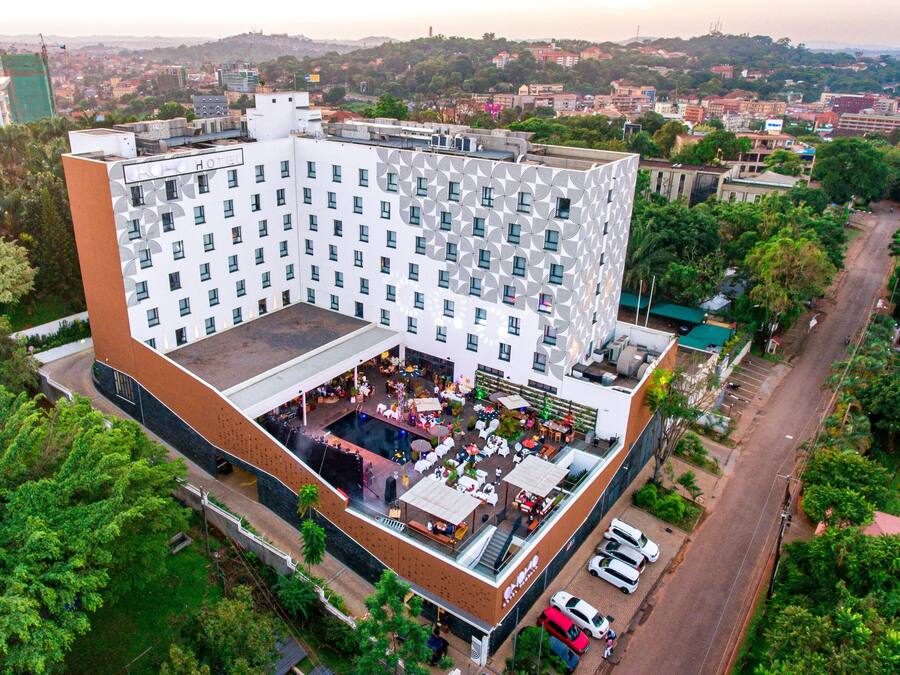 Vista aérea de un hotel con terraza en la azotea, rodeado de vegetación y un paisaje urbano de fondo.