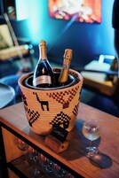 Una cesta tejida con dos botellas de champán, una copa de champán y un posavasos de madera.