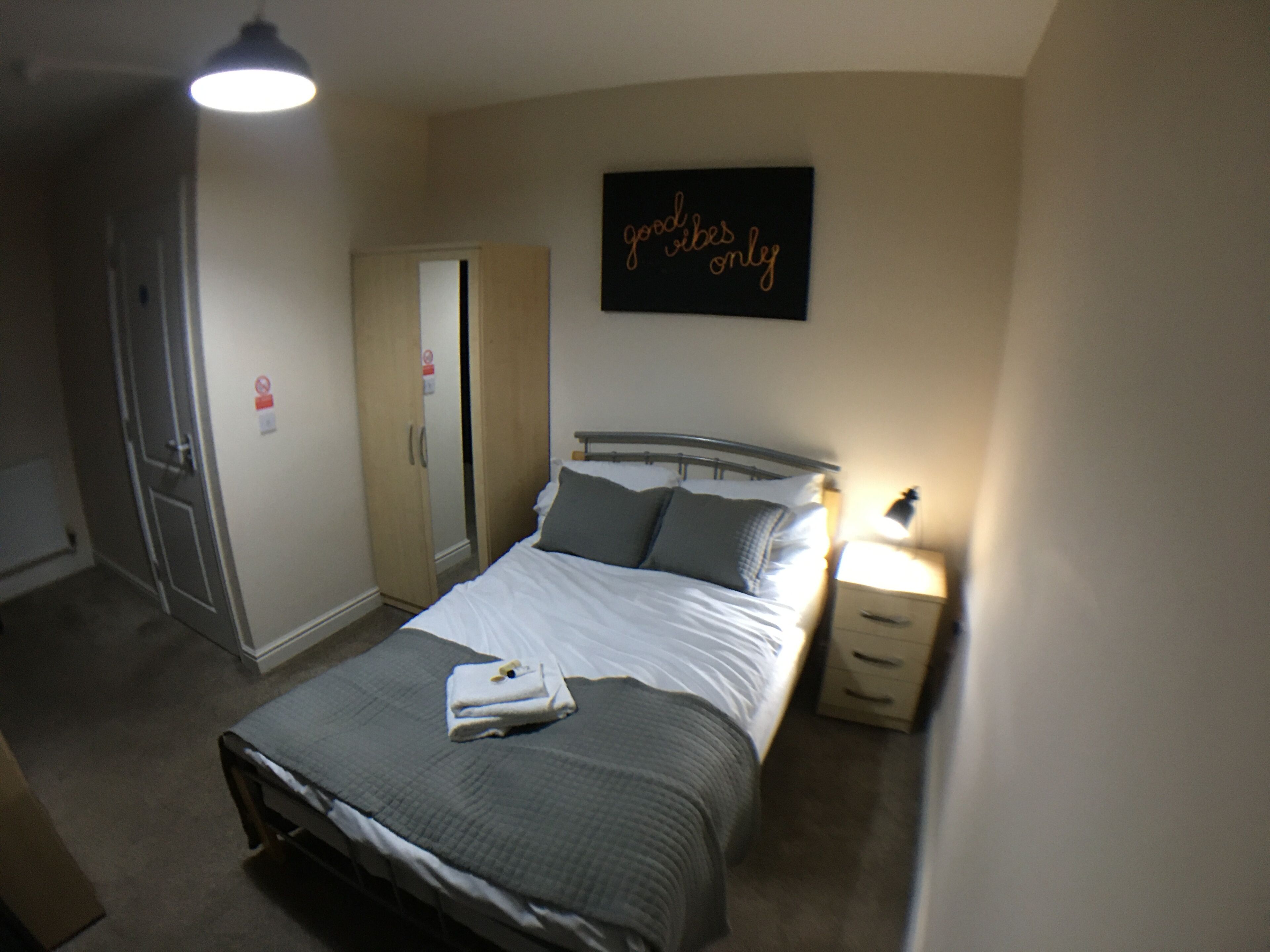 A bedroom with a bed, bedside table, nightstand, wardrobe, and a wall sign.
