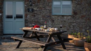 A stone building with a wooden picnic table set up outside, featuring a wine bottle, bowls, and bread. - Barn Conversion With Indoor Swimming Pool In Stunning Rural Location Near Ocean (Bude)