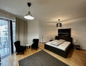 Quarto moderno com uma cama grande, uma escrivaninha, duas cadeiras, um abajur e vista para a cidade pela janela.