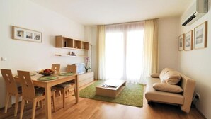 Ein modernes Wohnzimmer mit einem Holzesstisch, Stühlen, einer Couch und einem kleinen Couchtisch. Über dem Tisch befinden sich Regale und ein Fenster mit Vorhängen.