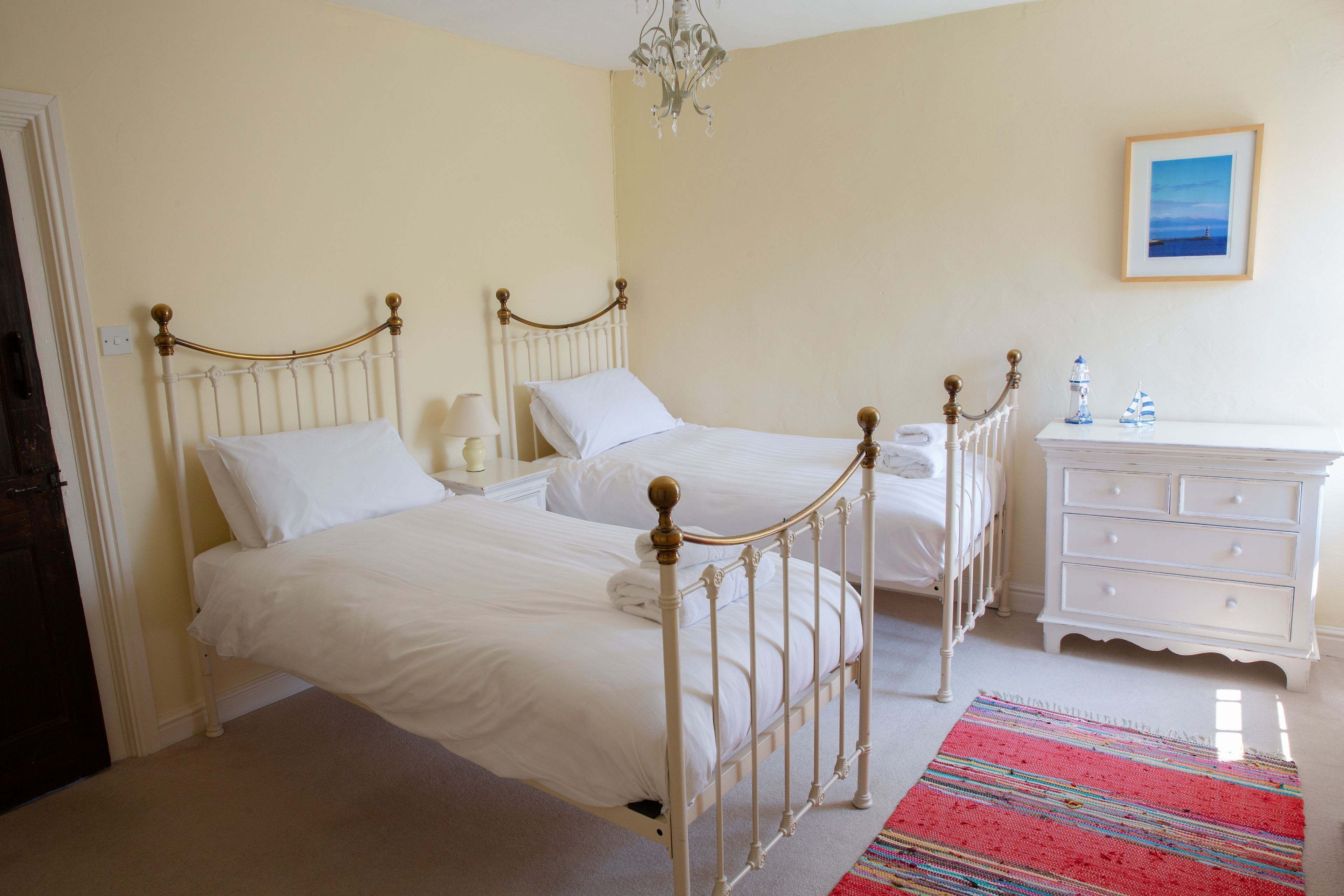 A bedroom with two single beds, a white dresser, a chandelier, and a framed picture on the wall.