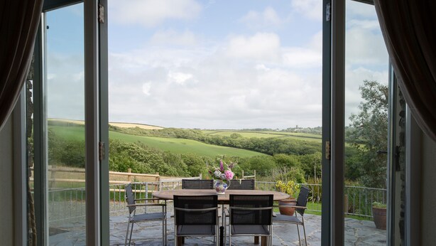 A patio with a stone table and chairs, a vase with flowers, and a view of green hills. - Luxury Cottage With Indoor Swimming Pool In Stunning Rural Location Near Ocean (Bude)