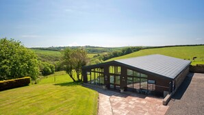 A modern building with large windows and a sloped roof, surrounded by a well-maintained lawn and trees. - Luxury Cottage With Indoor Swimming Pool In Stunning Rural Location Near Ocean (Bude)