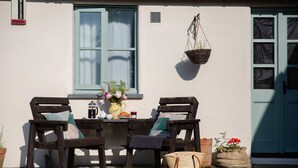 En rustik utomhusplats med två trästolar, ett litet bord med en kaffekanna och en hängkorg med blommor.