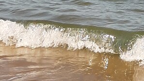 Blick auf die Küste mit an die Ufer treibenden Wellen und einem Gebäude in der Ferne auf dem Wasser.