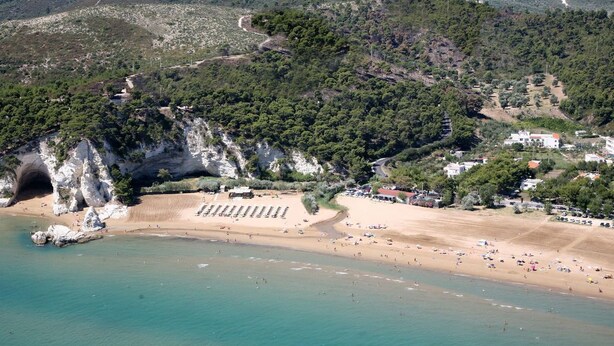 Vue aérienne d’une zone côtière avec une plage, des falaises rocheuses et un paysage montagneux en arrière-plan.