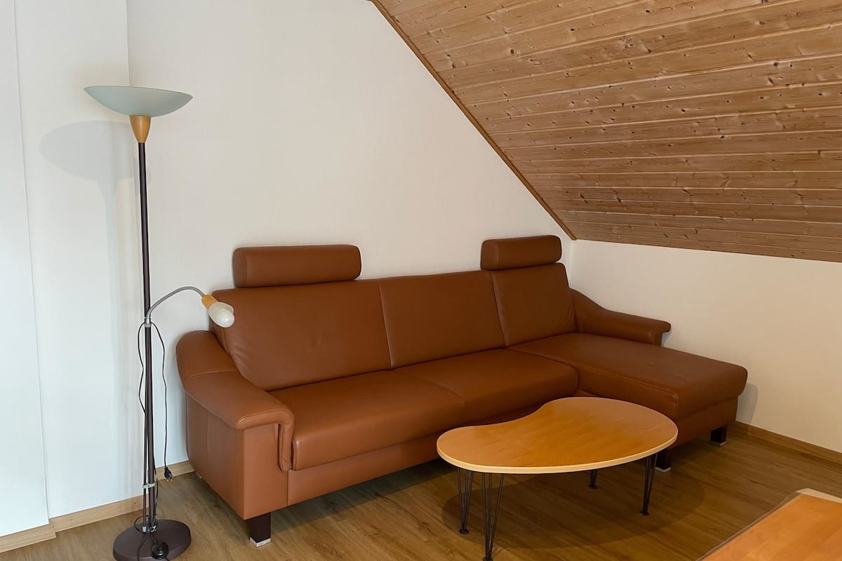 A brown leather sofa, a small wooden table, and a floor lamp in a room with wooden flooring and a sloped ceiling.