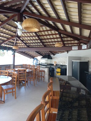 Un espace repas extérieur avec des chaises et des tables en bois, un coin cuisine équipé d’un réfrigérateur et d’une cuisinière, et un plafond orné de poutres en bois et de luminaires tressés.