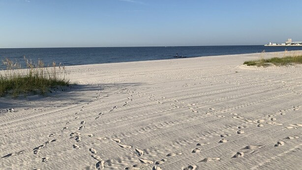 Una playa arenosa con huellas que se dirigen al agua, cielo despejado y un edificio a lo lejos.