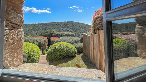 Blick durch ein Fenster mit steinernen Bögen, einem Holzzaun und einem gepflegten Garten mit verschiedenen Sträuchern und einer Hügelkulisse im Hintergrund.