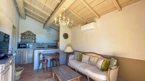 Ein gemütliches Wohnzimmer mit einer Couch, einem Holztisch und einem Essbereich mit Tisch und Stühlen. Im Hintergrund ist eine Küche erkennbar, darin ein Mikrowellenherd und eine Wanduhr.