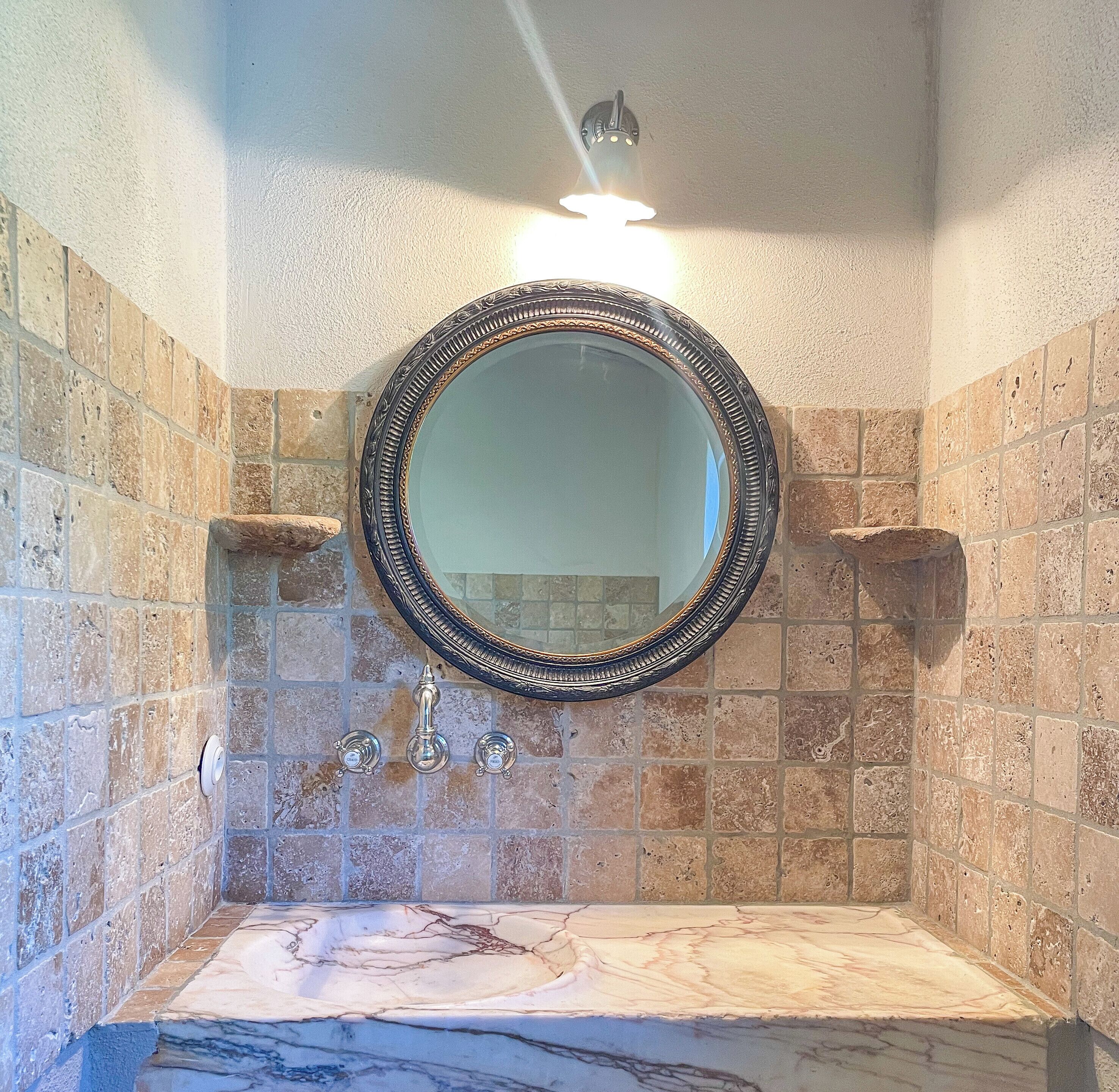 Un baño con un lavamanos de mármol, un espejo redondo con un marco ornamentado y una pared con azulejos de piedra.