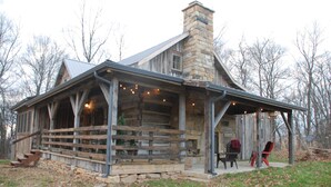 A rustic log cabin with a stone chimney, a covered porch, and a seating area with chairs.