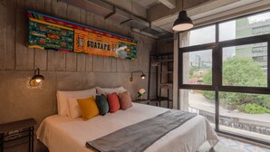 Een slaapkamer met een groot bed, een kleurrijke wanddecoratie, een raam met uitzicht op gebouwen en bomen, en een klein tafeltje met een lamp.