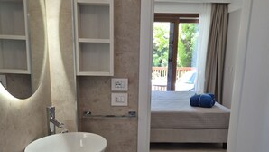 Ein modernes Badezimmer mit einem großen, ovalen Waschbecken, einem Spiegel und einer Holzausstattung. Durch einen offenen Durchgang ist ein Bett sichtbar, das auf einen angrenzenden Schlafbereich hindeutet.