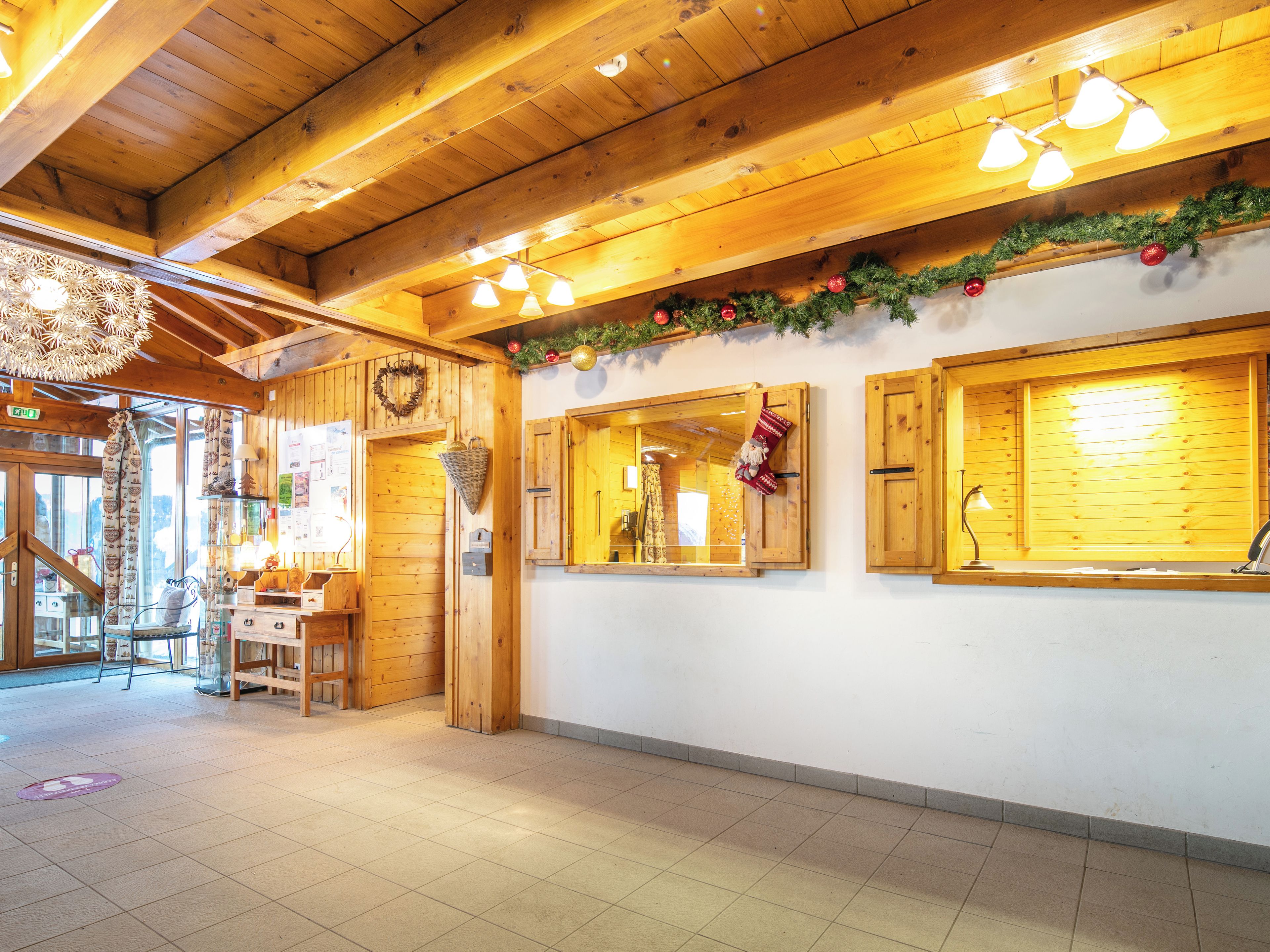A wooden interior with a high ceiling, wooden beams, and a tiled floor. There are windows with wooden shutters, a wall-mounted light fixture, and a Christmas garland.