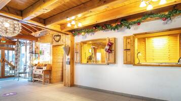 A wooden interior with a high ceiling, wooden beams, and a tiled floor. There are windows with wooden shutters, a wall-mounted light fixture, and a Christmas garland.