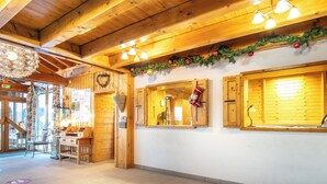 Een houten interieur met een hoge plafond, houten balken en een betegelde vloer. Er zijn ramen met houten luiken, een wandlamp en een kerstkrans.
