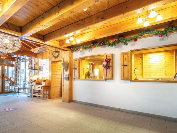 A wooden interior with a high ceiling, wooden beams, and a tiled floor. There are windows with wooden shutters, a wall-mounted light fixture, and a Christmas garland. - Residence La Duit - Vacanceole (La Léchère)
