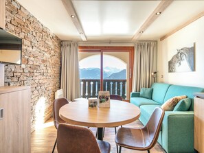 Una habitación acogedora con pared de piedra, techo de madera, una mesa redonda con sillas, un sofá verde y vistas a montañas a través de un gran ventanal.