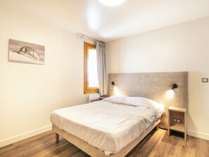 A bedroom with a bed, a nightstand, a wall-mounted lamp, a window with curtains, and a painting on the wall. - Residence La Duit - Vacanceole (La Léchère)