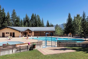 A pool area with a building, wooden deck, and surrounding trees. - Residence La Duit - Vacanceole (La Léchère)