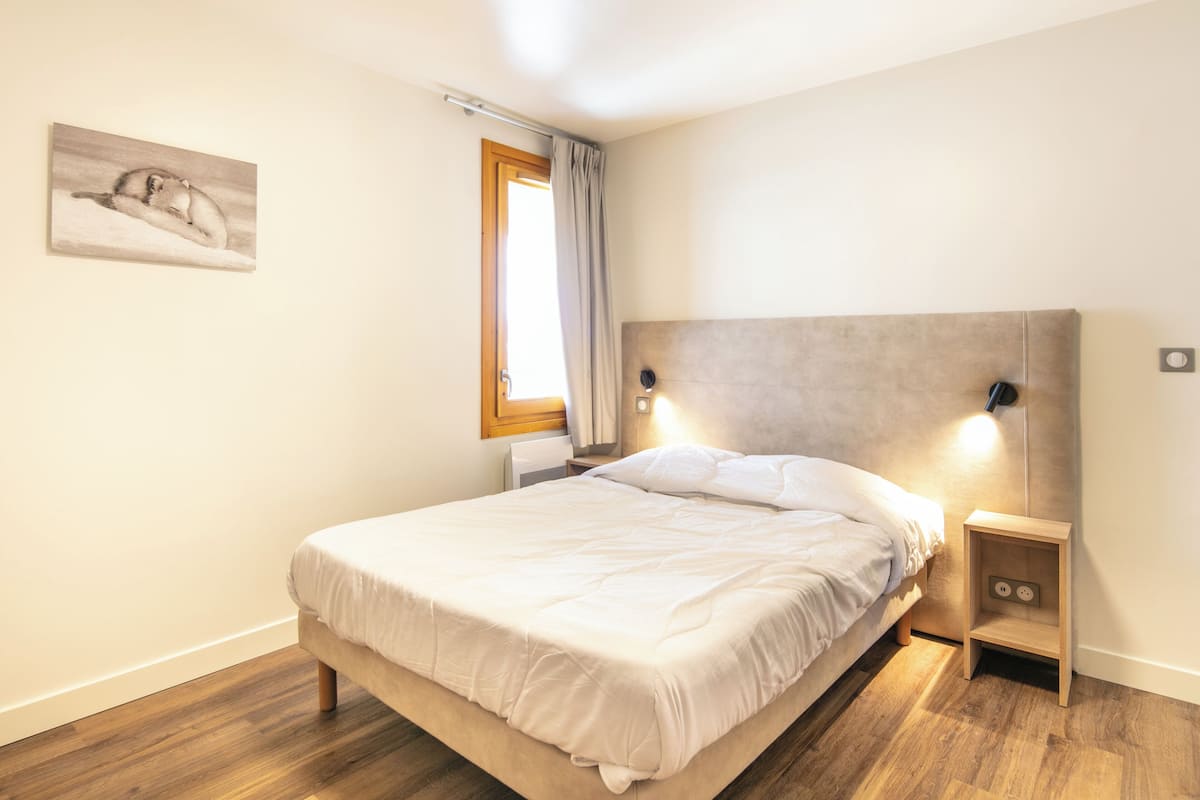 A bedroom with a bed, a nightstand, a wall-mounted lamp, a window with curtains, and a painting on the wall.