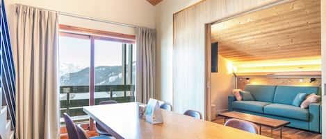 A modern room with a wooden ceiling, a large table with chairs, a sofa, and a view of snow-capped mountains through large windows.