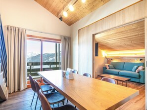 Una habitación moderna con techo de madera, una mesa grande con sillas, un sofá y vistas a montañas nevadas a través de amplios ventanales.