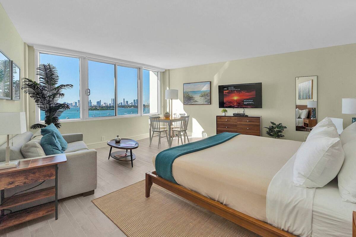 A modern hotel room with a large bed, a desk, a chair, a TV, and a view of the city skyline.