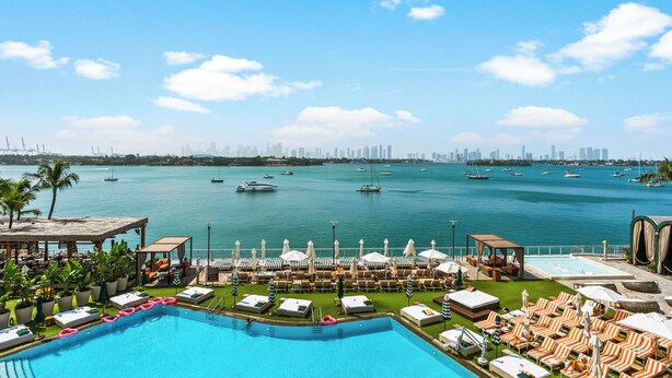 A poolside area with lounge chairs and umbrellas, a clear view of the water, and a city skyline in the distance.