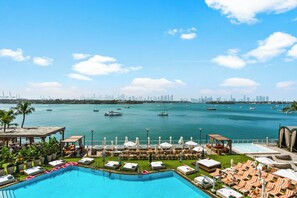 Un área junto a la piscina con sillas de descanso y sombrillas, una vista despejada del agua y el horizonte de una ciudad a lo lejos.