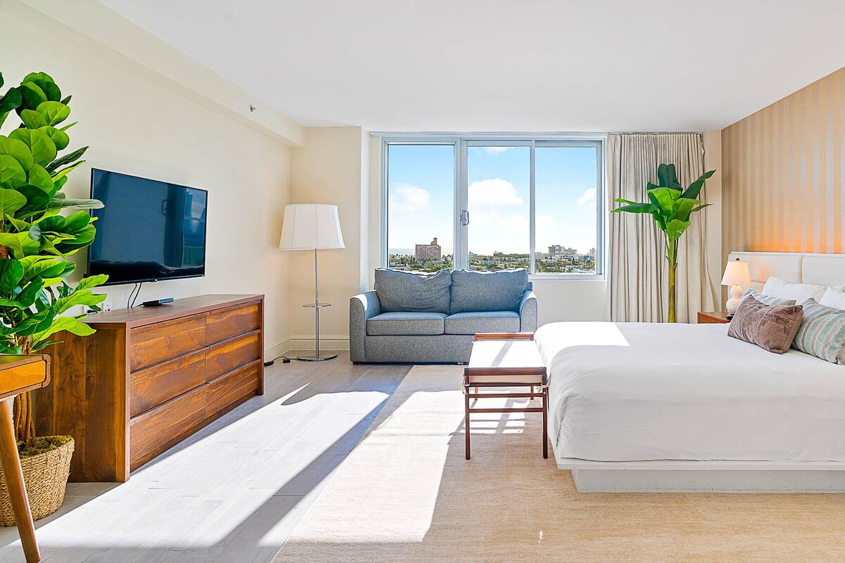 A modern hotel room with a large bed, a sofa, a TV, and a view of the city.