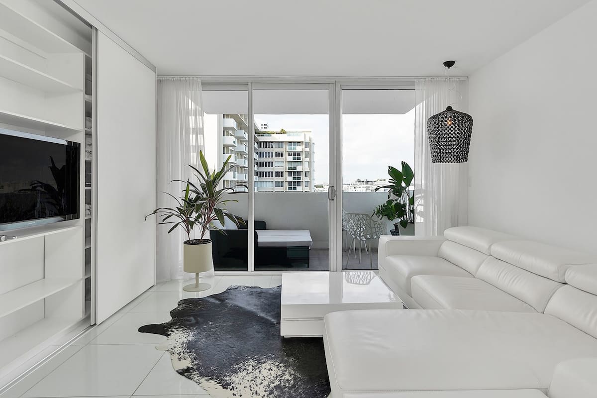 A modern living room with a white sofa, a flat-screen TV, and a large glass door leading to a balcony with a view of a building.