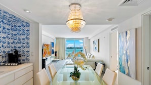 A modern dining room with a glass table, white chairs, a chandelier, and a view of the ocean through large windows.