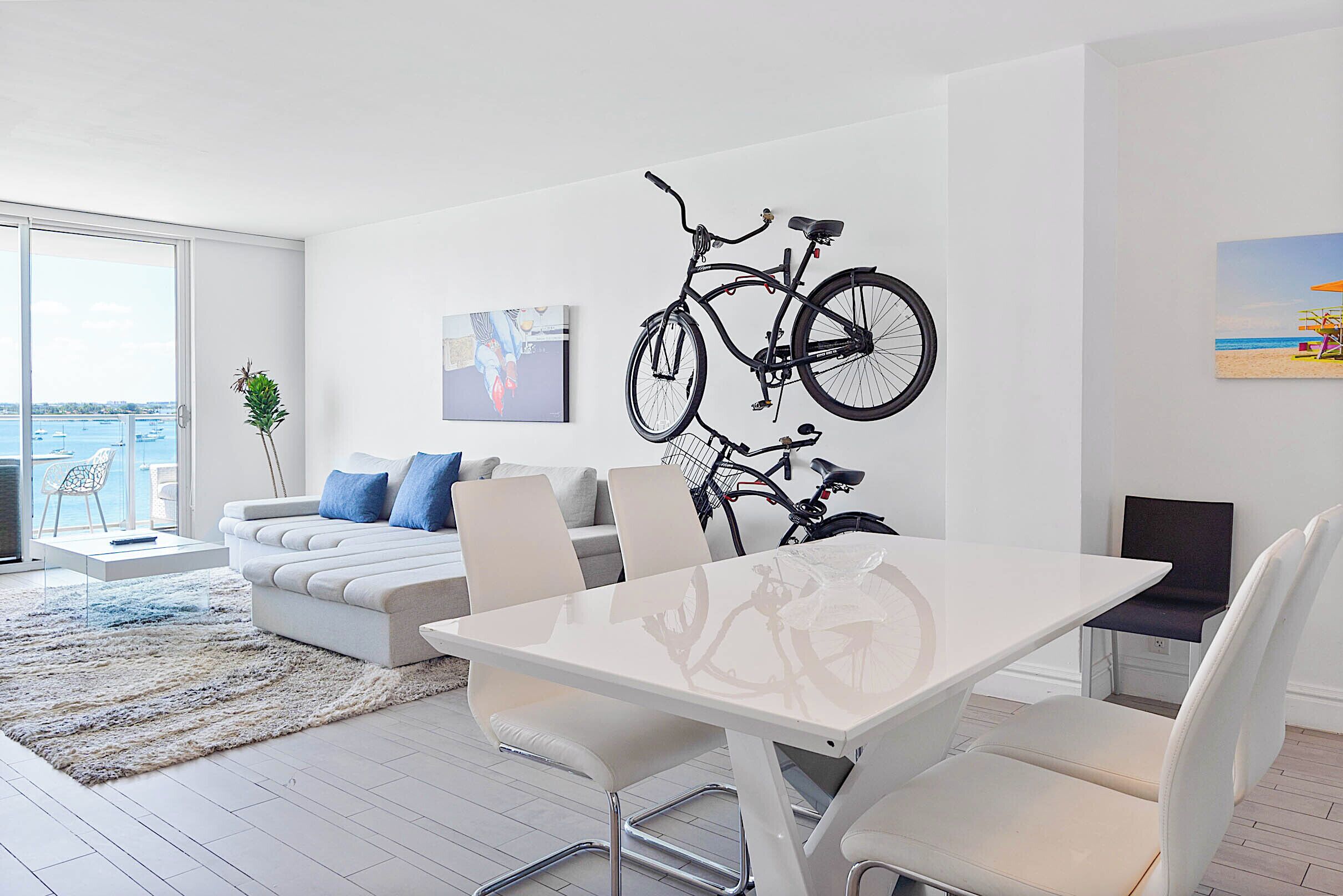 A modern living room with a white sofa, a dining table, and a large window overlooking the sea.