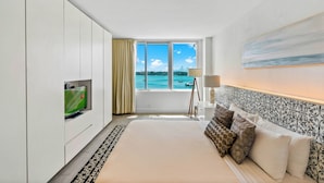 A hotel room with a large bed, a television on a cabinet, a window with a view of the sea, and a patterned headboard.