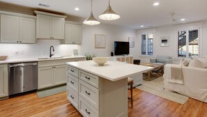 A modern kitchen with white cabinets, a stainless steel dishwasher, and a central island with a bowl. There is a dining area with a table and chairs, and a living room with a sofa and a TV.