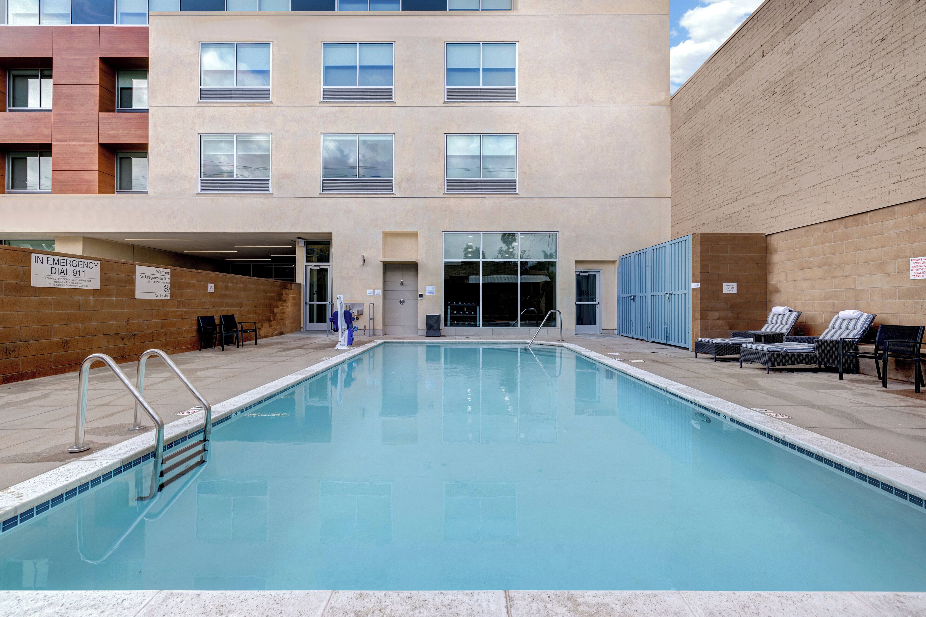 Una piscina con un puesto de salvavidas, sillas de descanso y un edificio con múltiples ventanas al fondo.