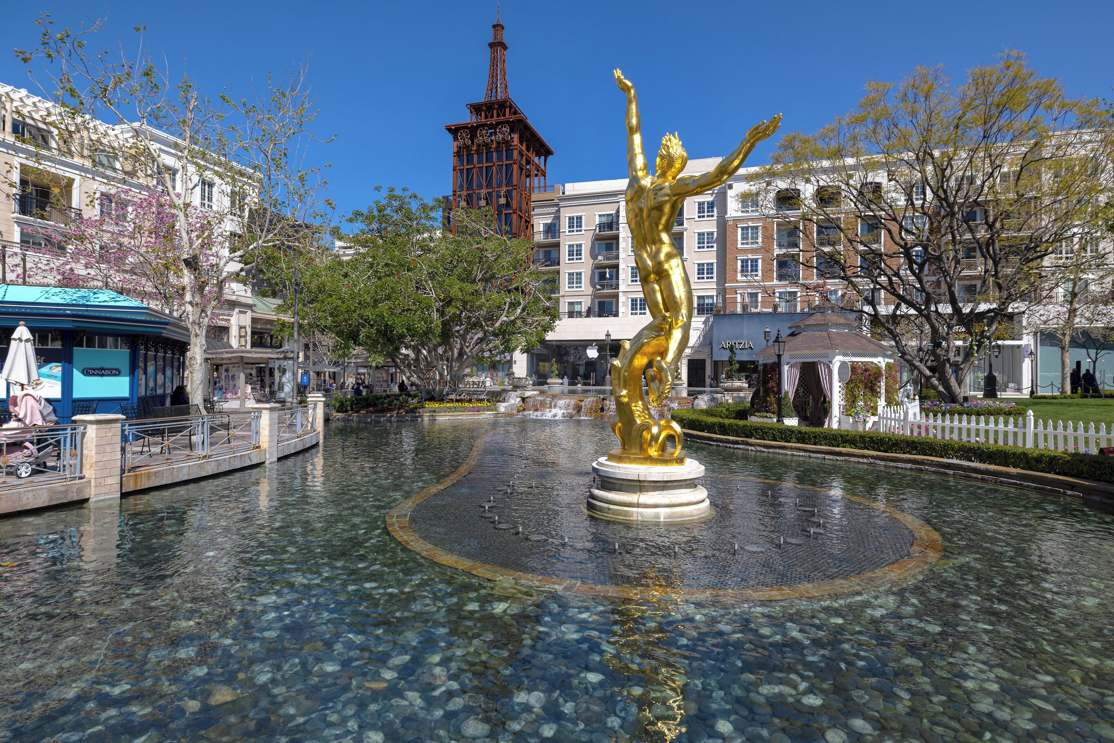 Uma fonte de água cristalina com uma estátua dourada no centro, rodeada por um espaço pavimentado com bancos e um edifício ao fundo.