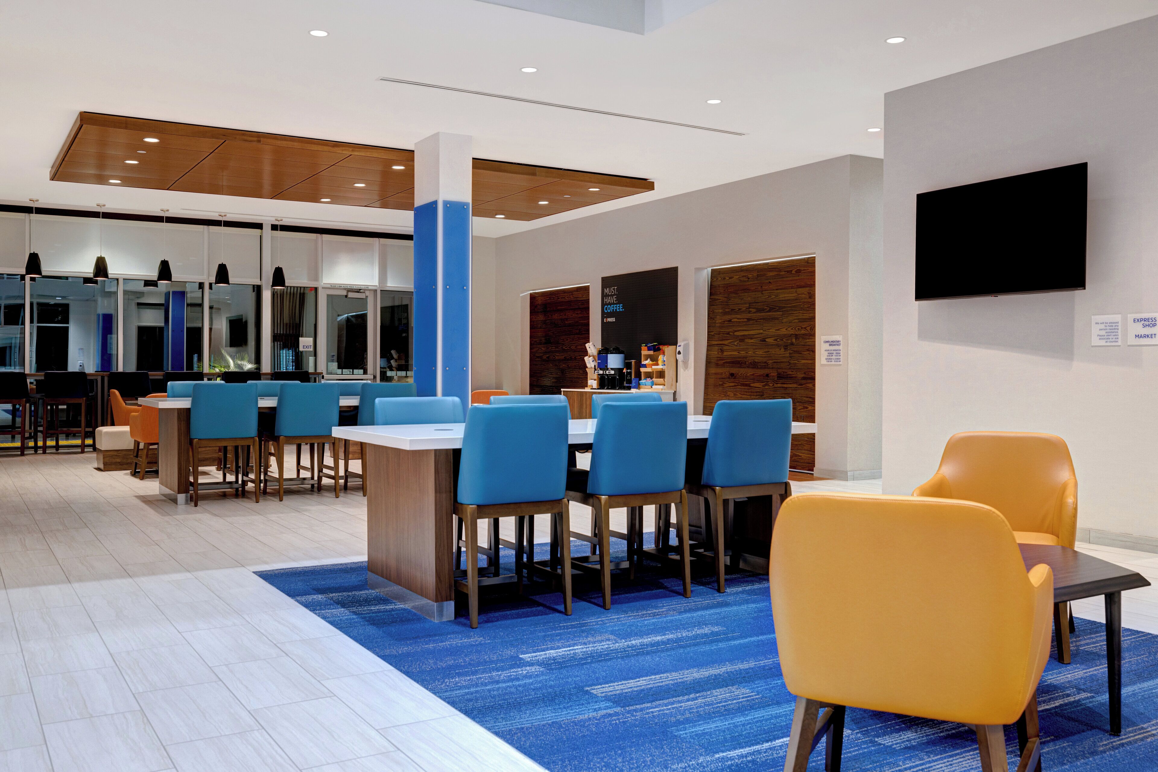A modern dining area with blue and yellow chairs, a coffee machine, and a flat-screen TV on the wall.