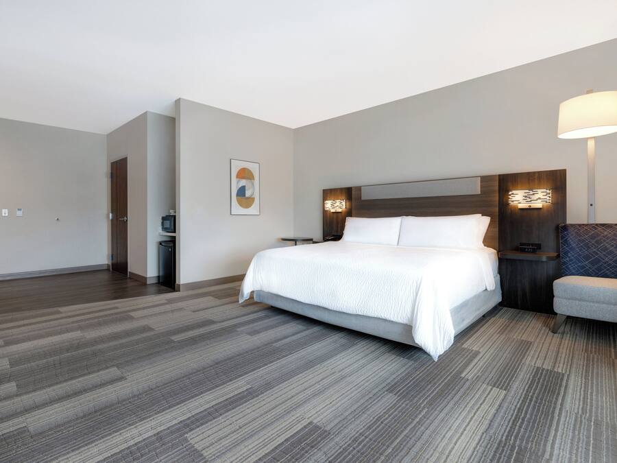 A hotel room with a bed, a chair, a nightstand, a wall-mounted lamp, and a door.
