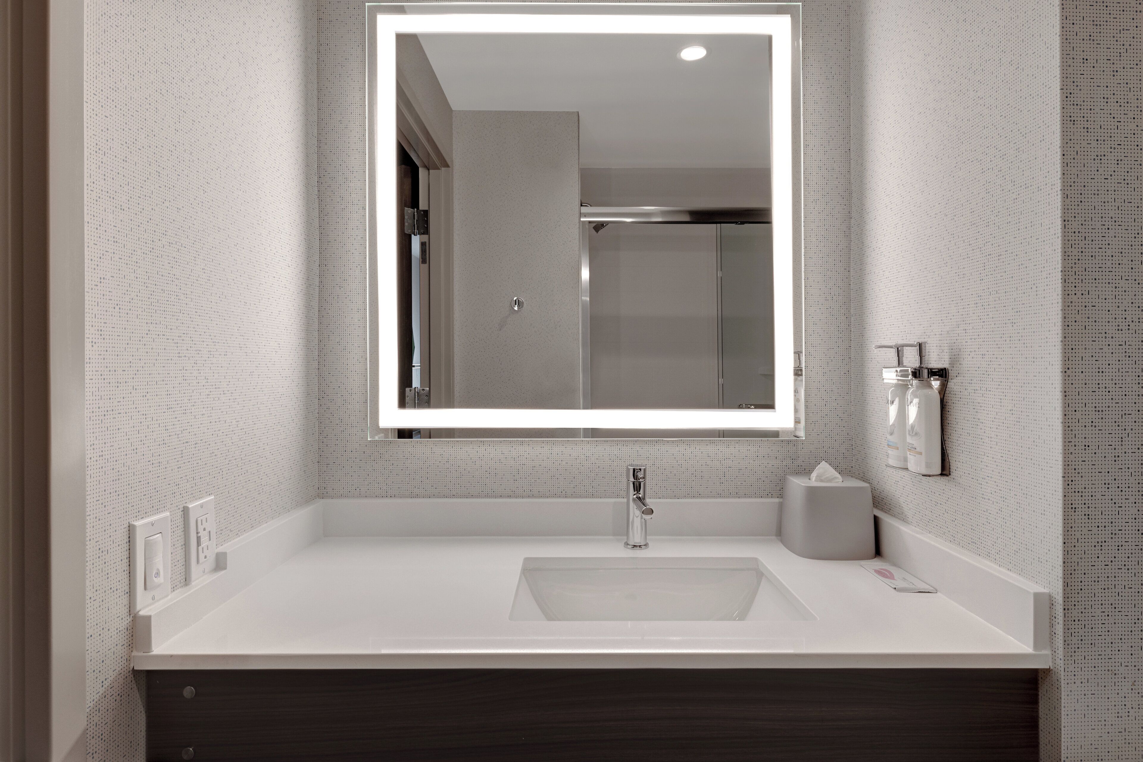 Un baño con lavamanos blanco, un espejo con luz y un dispensador de toallas.