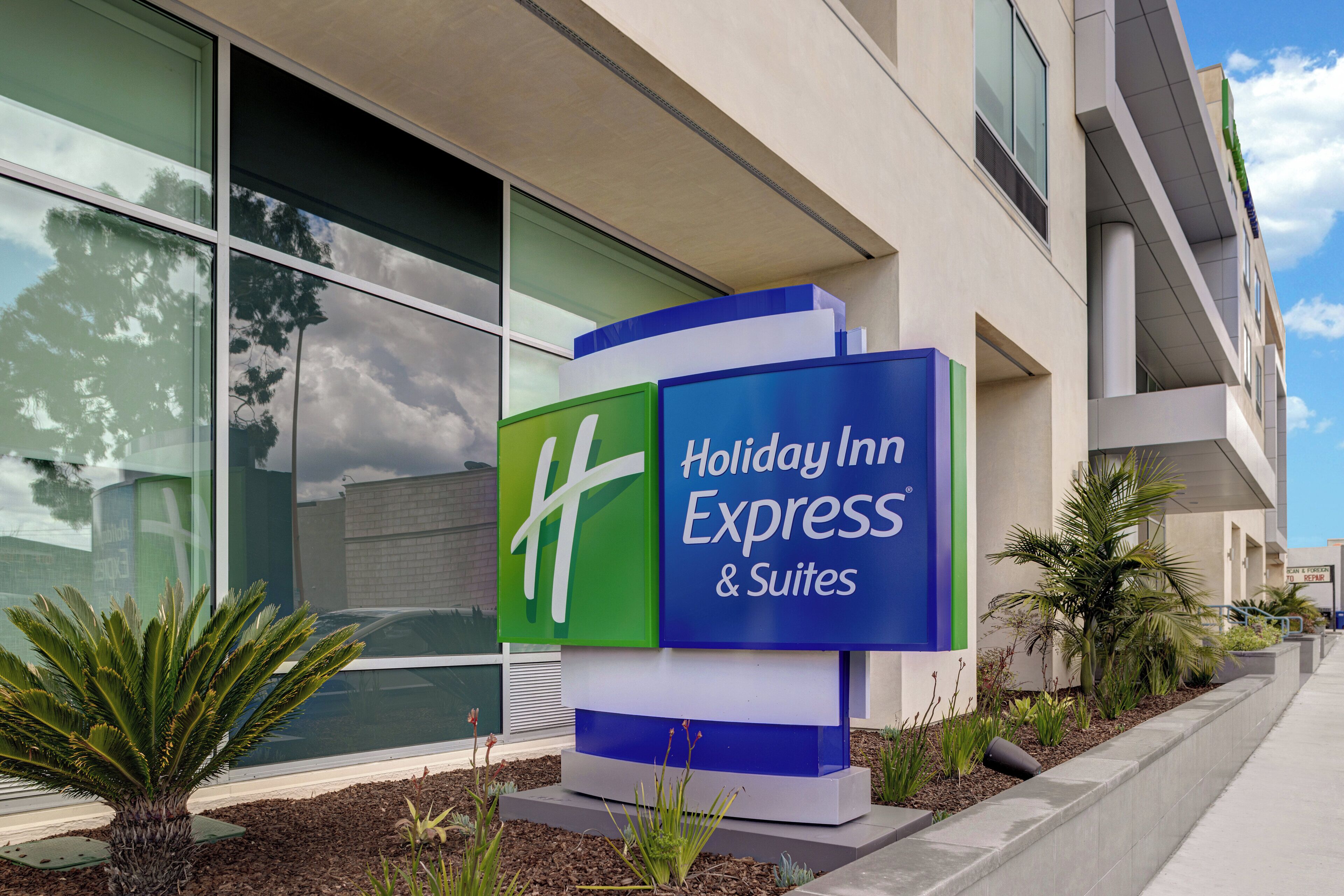 A Holiday Inn Express & Suites sign with a modern design, located in front of a building with large windows and a clear sky.