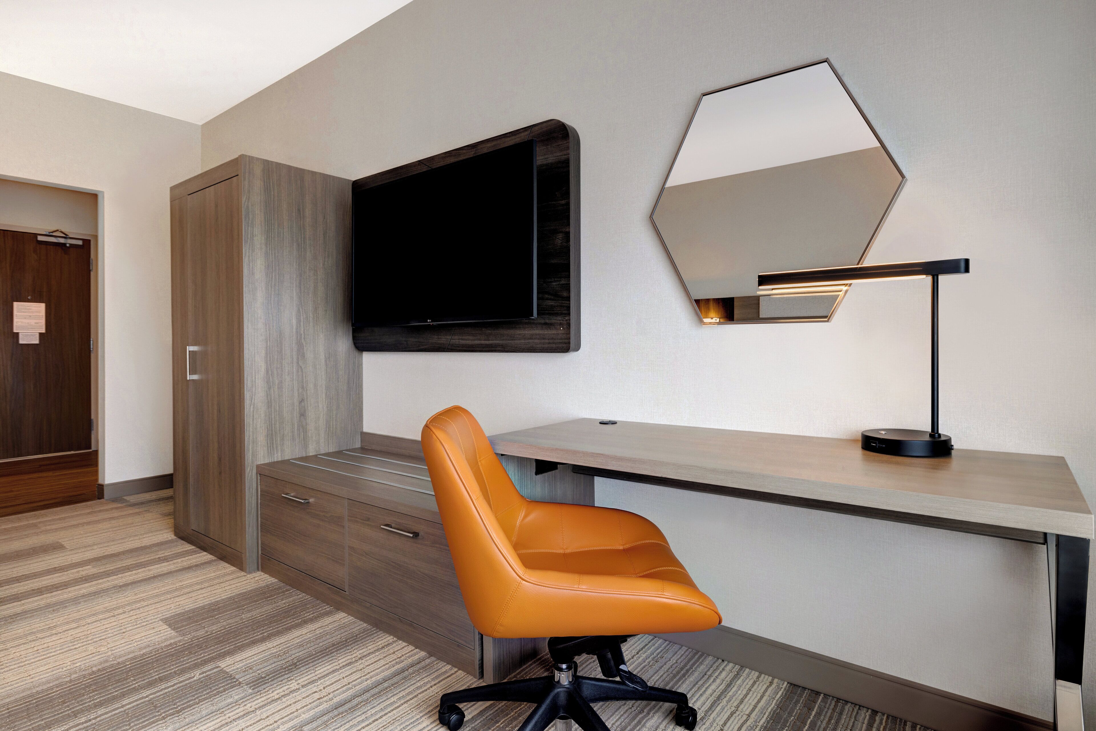 Un espacio de oficina moderno con un escritorio, una silla de oficina naranja, un televisor de pantalla plana, un espejo montado en la pared y una lámpara de escritorio.