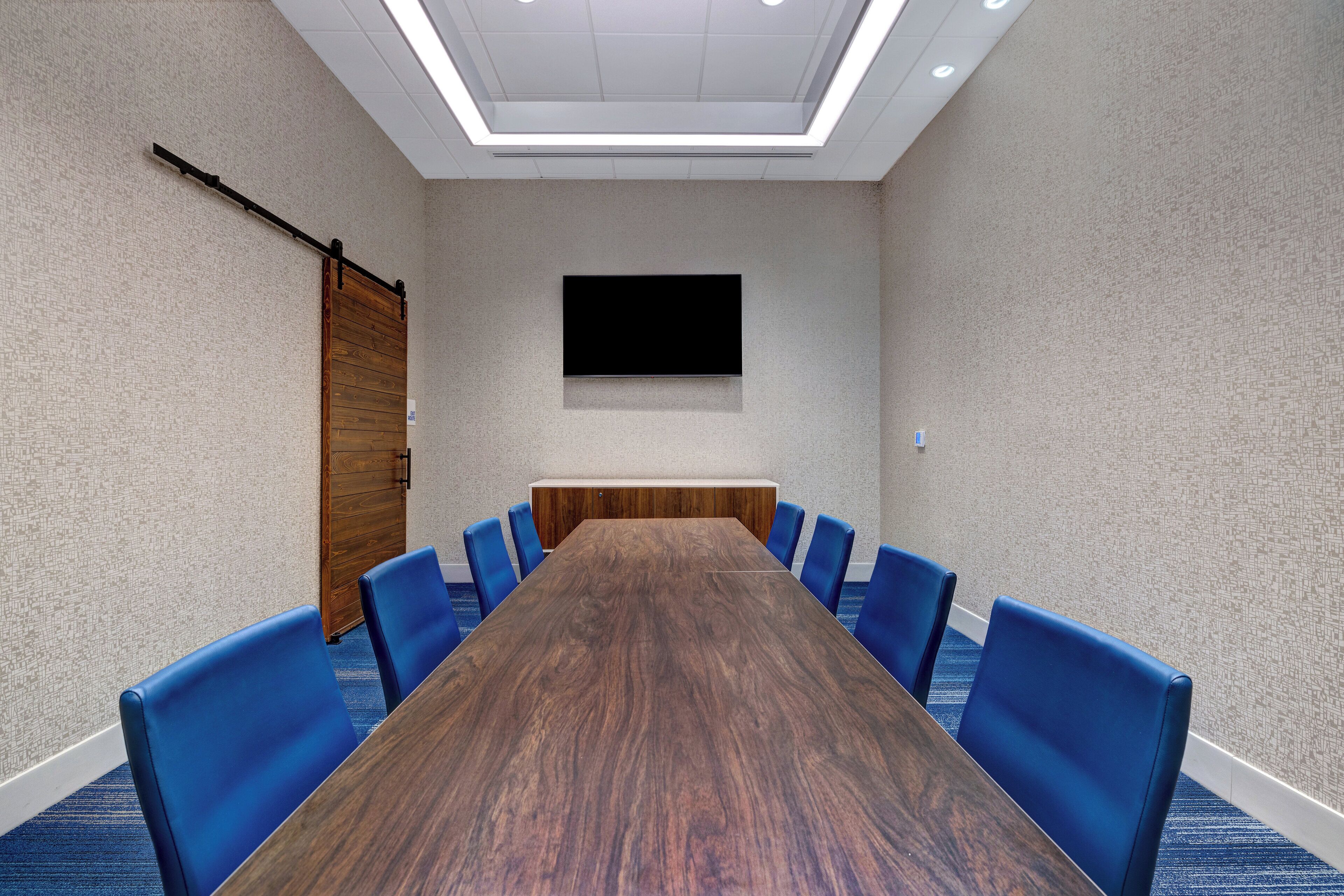 Une salle de conférence avec une longue table en bois, des chaises bleues, un téléviseur à écran plat fixé au mur et un luminaire au plafond.